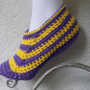 Puede incluir: Pantufla de crochet a rayas moradas y amarillas. La pantufla está hecha con un hilo suave y tiene un ajuste cómodo.