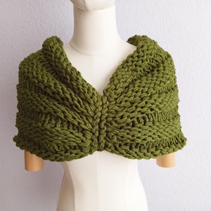 Puede incluir: Chal tejido de color verde oliva con diseño de cuello en V. El chal está hecho de hilo grueso y tiene una apariencia texturizada. La prenda se exhibe en un maniquí.