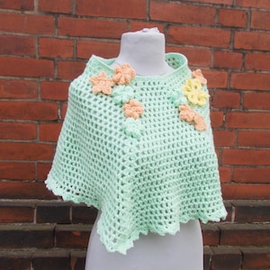 Könnte beinhalten: Ein hellgrünes gehäkeltes Poncho mit floralem Design. Der Poncho hat einen quadratischen Ausschnitt und ist mit pfirsichfarbenen und gelben gehäkelten Blumen verziert. Das Kleidungsstück wird auf einer Schaufensterpuppe vor einer Backsteinmauer präsentiert.
