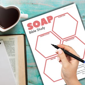 May include: A printable SOAP Bible study worksheet with a pink hexagon shape for each section: Scripture, Observation, Application, and Prayer. The worksheet is on a blue wooden surface.