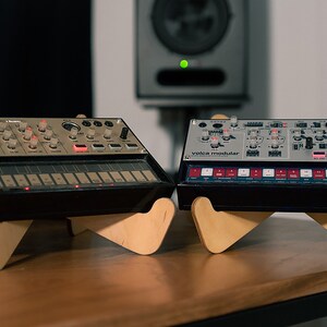 May include: Two small, black, electronic music synthesizers, a Korg Volca Modular and a Korg Volca Beats, are on wooden stands. The synthesizers have a variety of knobs, buttons, and lights. The Volca Modular has the text "volca modular" printed on the front.