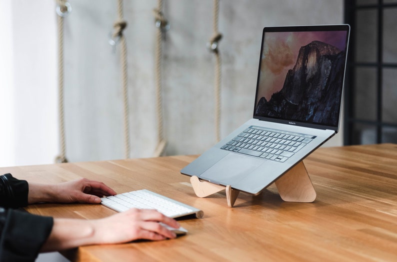 May include: A silver laptop computer is on a wooden laptop stand. The laptop is open and the screen shows a landscape image. A white wireless keyboard is on the table in front of the laptop.