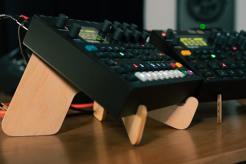 May include: Two black electronic music synthesizers with colorful buttons and knobs, sitting on wooden stands. The synthesizers are on a wooden table.