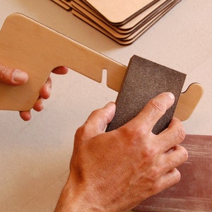 May include: A person sanding a wooden piece with a brown sanding block. The wooden piece has a cut-out shape and is being held in the left hand. The sanding block is being held in the right hand.