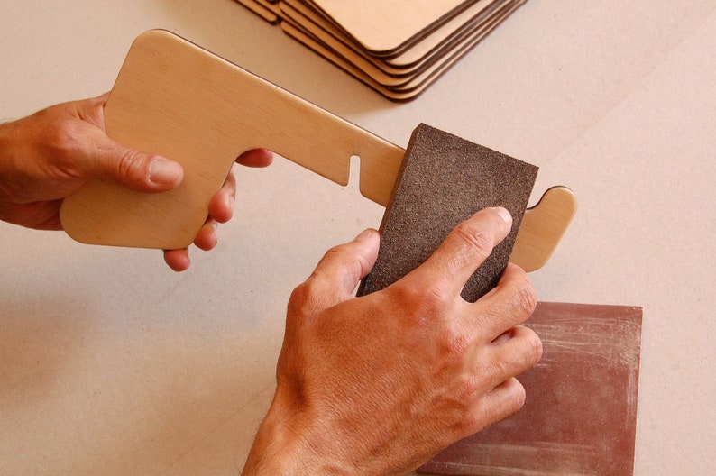 May include: A person sanding a wooden piece with a brown sanding block. The wooden piece has a cut-out shape and is being held in the left hand. The sanding block is being held in the right hand.