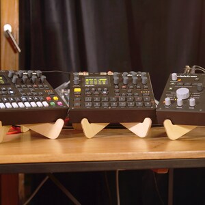 May include: Three black electronic music synthesizers on wooden stands. The synthesizers have various buttons, knobs, and a digital display. The synthesizers are on a wooden table.