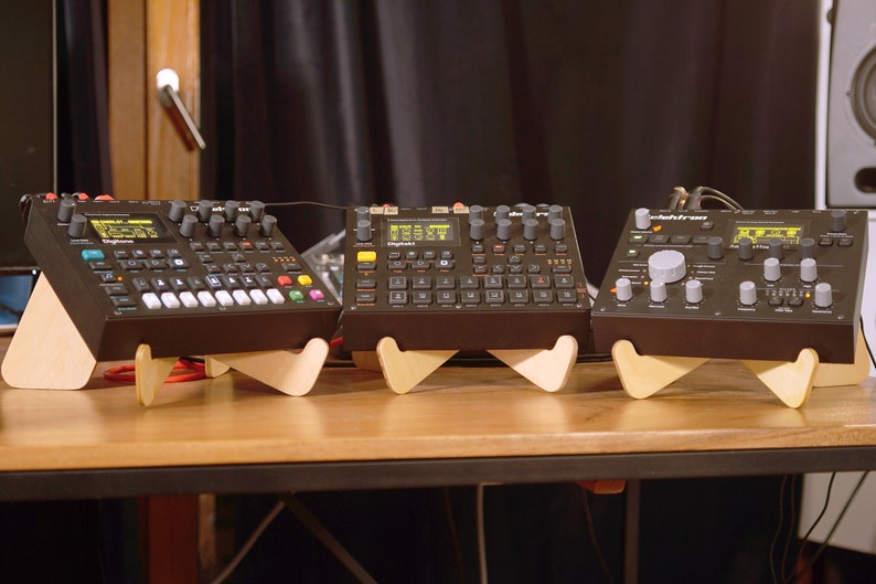 May include: Three black electronic music synthesizers on wooden stands. The synthesizers have various buttons, knobs, and a digital display. The synthesizers are on a wooden table.
