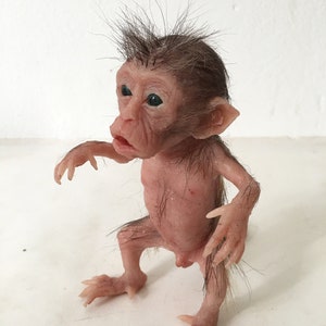 May include: A small, realistic-looking monkey doll made of silicone. The doll has brown hair, green eyes, and a pink body. It is standing on a white surface.