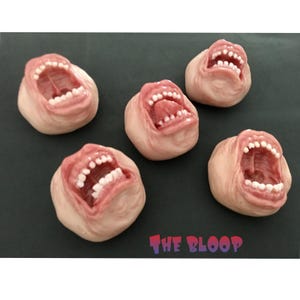 Op de afbeelding: Vijf keramische sculpturen van open monden met tanden, elk met een andere uitdrukking. De sculpturen zijn geschilderd in tinten roze en wit en zijn gerangschikt op een donkergrijs oppervlak. De tekst "THE BLOOP" is zichtbaar in de rechterbenedenhoek van de afbeelding.
