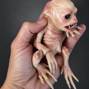 May include: A small, pale, flesh-toned creature figurine with wrinkled skin, a large head, dark eyes, and a mouth filled with sharp, pointed teeth. The creature has long, slender limbs with claw-like fingers and toes, and a textured, organic appearance.