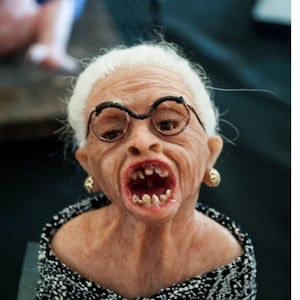 Könnte beinhalten: Eine Keramikbüste einer älteren Frau mit weißen Haaren, die eine Brille und ein schwarz-weiß gemustertes Kleid trägt. Ihr Mund ist offen und sie zeigt ihre Zähne.