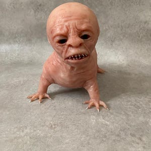 Puede incluir: Escultura de criatura reptante de tono rosado, con rostro arrugado, ojos pequeños y dientes afilados. La escultura tiene manos y pies con forma de garra, sobre un fondo neutro.