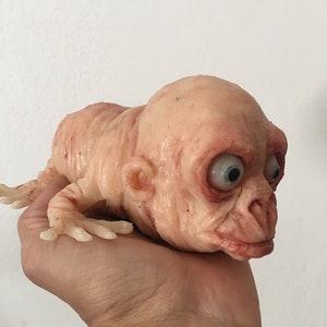 May include: A small, fleshy, humanoid figure with large eyes and a wrinkled face. The figure is made of a soft, rubbery material and is painted in shades of pink and red.