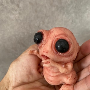 Könnte beinhalten: Kleine, fleischige Skulptur einer Kreatur mit großen schwarzen Augen und einem blassrosa Hautton. Die Figur hat kleine Arme und Beine und wird in einer Hand gehalten. Die Skulptur hat ein detailliertes, strukturiertes Aussehen.