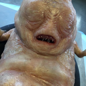 Puede incluir: Primer plano de una figura humanoide pálida y carnosa con una cabeza grande y una boca ancha y llena de dientes. La figura tiene una superficie arrugada y texturizada y parece estar hecha de un material blando y maleable.