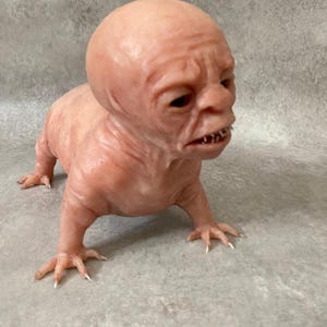 May include: A detailed sculpture of a creature with a bald head, wrinkled skin, and clawed hands and feet. The figure is a fleshy pink color and is posed on all fours.