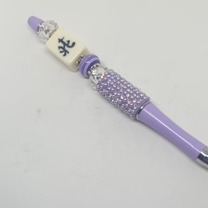May include: A lavender ballpoint pen featuring a decorative topper with a clear faceted bead, a cream-colored rectangular mahjong tile with a black character, a purple and white striped bead, and a cylindrical section covered in lavender rhinestones.