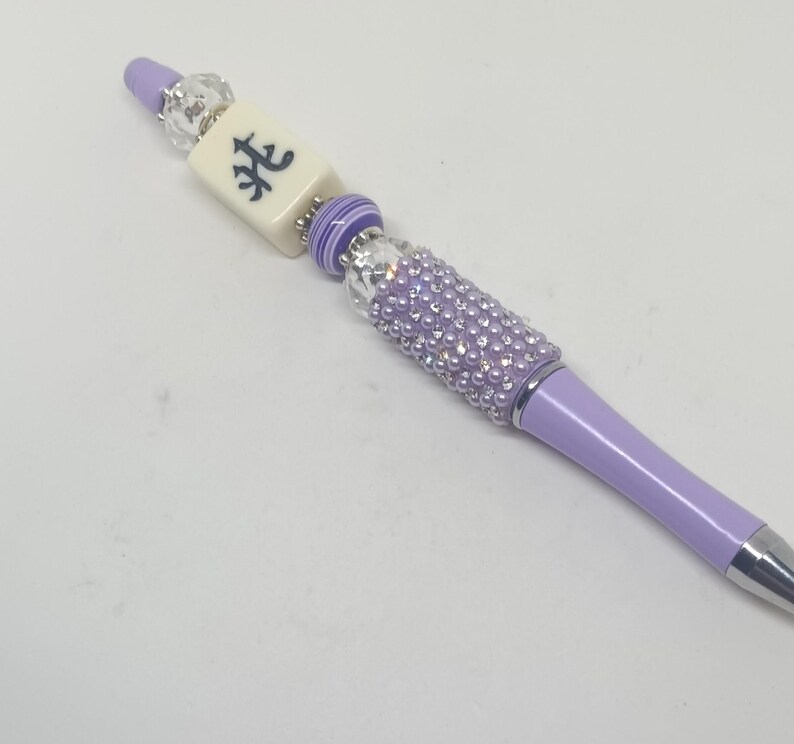 May include: A lavender ballpoint pen featuring a decorative topper with a clear faceted bead, a cream-colored rectangular mahjong tile with a black character, a purple and white striped bead, and a cylindrical section covered in lavender rhinestones.