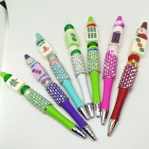 May include: Six ballpoint pens featuring Mahjong tile toppers and barrels embellished with colorful rhinestones. Each pen displays a unique combination of vibrant plastic colors, including lime green, purple, teal, white, and hot pink, with metallic tips.