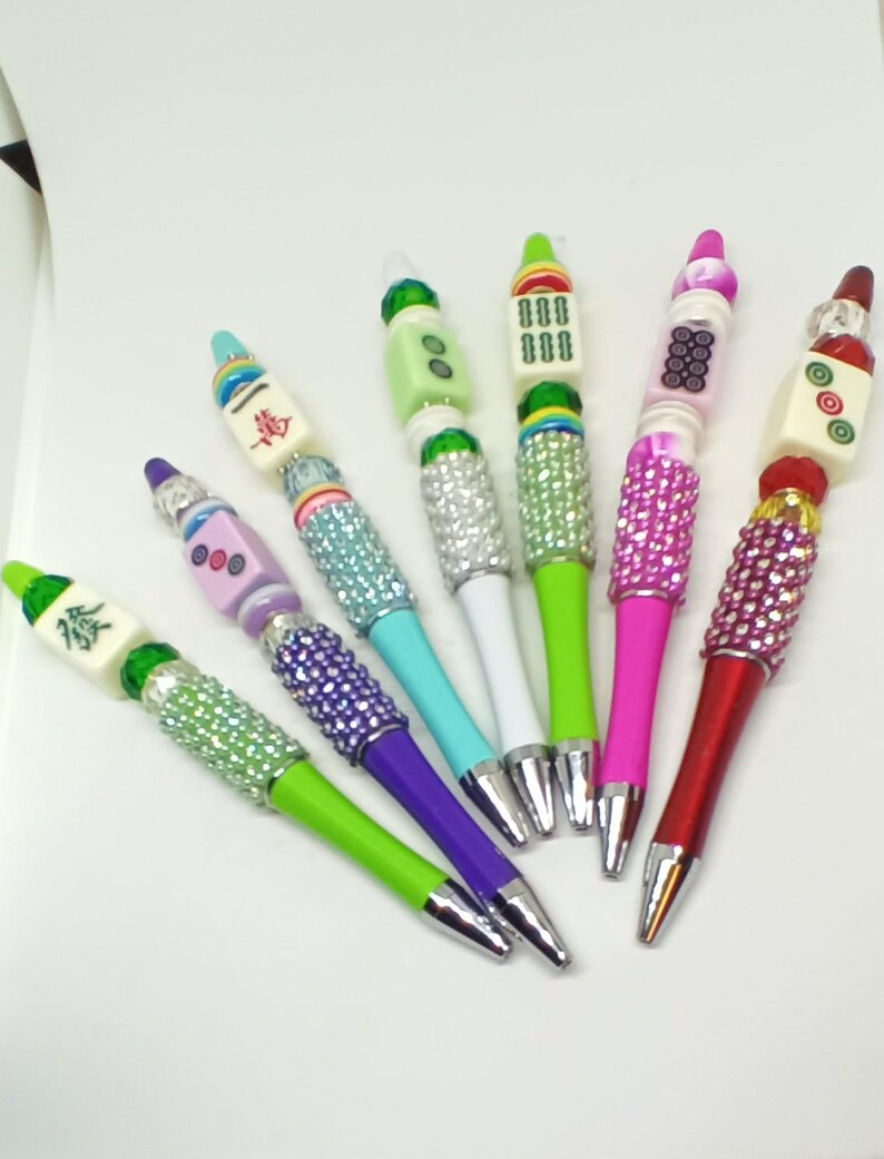May include: Six ballpoint pens featuring Mahjong tile toppers and barrels embellished with colorful rhinestones. Each pen displays a unique combination of vibrant plastic colors, including lime green, purple, teal, white, and hot pink, with metallic tips.