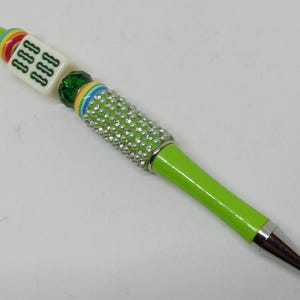 May include: A lime green ballpoint pen featuring a silver metal tip and a barrel adorned with clear rhinestones. The top section includes a multi-colored striped ring, a green faceted bead, and a white square mahjong tile with green symbols.