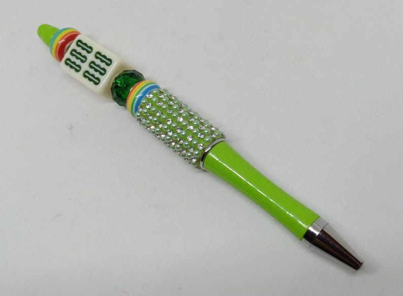 May include: A lime green ballpoint pen featuring a silver metal tip and a barrel adorned with clear rhinestones. The top section includes a multi-colored striped ring, a green faceted bead, and a white square mahjong tile with green symbols.