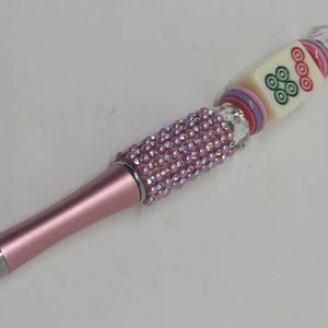 May include: A pink ballpoint pen featuring a barrel covered in small pink rhinestones and clear crystals. The upper section includes a square mahjong tile with green and red circular symbols, accented by stacked colorful rings and a clear bead topper.
