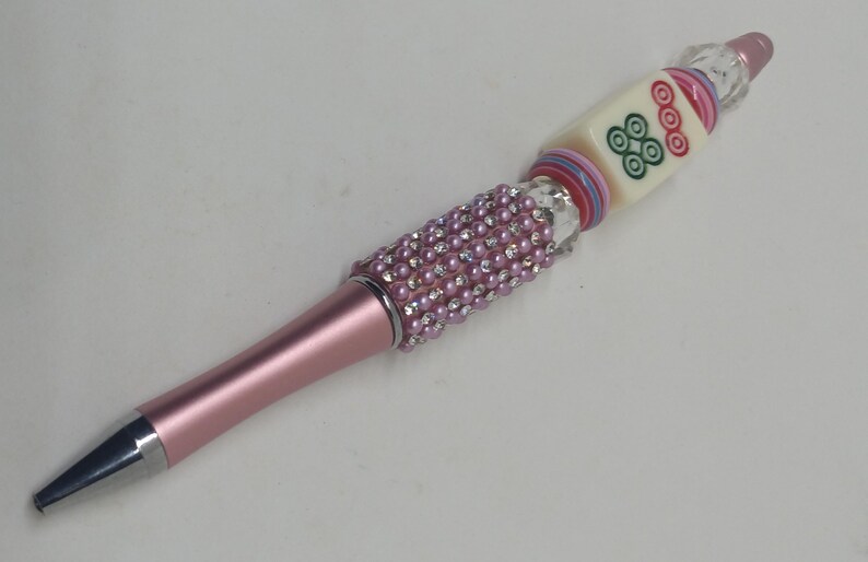 May include: A pink ballpoint pen featuring a barrel covered in small pink rhinestones and clear crystals. The upper section includes a square mahjong tile with green and red circular symbols, accented by stacked colorful rings and a clear bead topper.