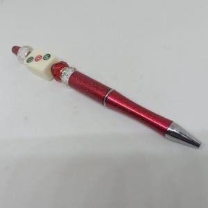 May include: A red glittery ballpoint pen featuring a decorative topper. The topper consists of a cream-colored square tile with three circular symbols in green and red, flanked by clear faceted beads and a red faceted bead at the base.