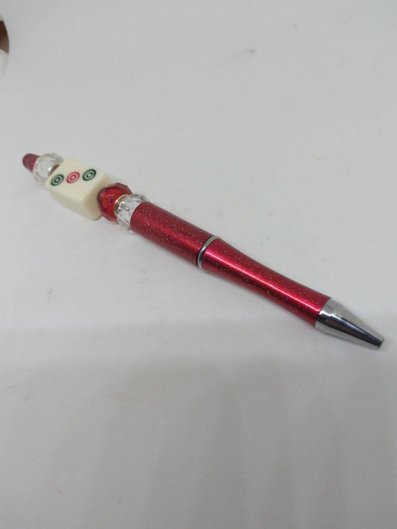May include: A red glittery ballpoint pen featuring a decorative topper. The topper consists of a cream-colored square tile with three circular symbols in green and red, flanked by clear faceted beads and a red faceted bead at the base.
