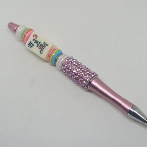 May include: A pink ballpoint pen featuring a decorative barrel adorned with light pink pearls and clear rhinestones. The upper section includes a white mahjong tile with a red floral design and the number 3, flanked by colorful striped beads.