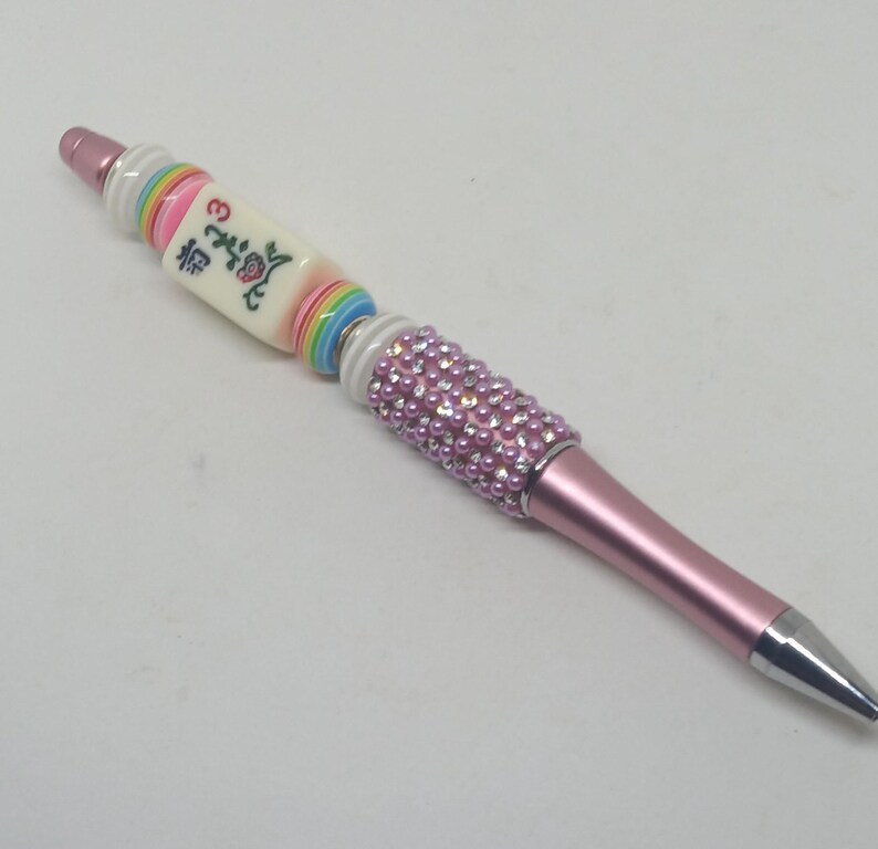 May include: A pink ballpoint pen featuring a decorative barrel adorned with light pink pearls and clear rhinestones. The upper section includes a white mahjong tile with a red floral design and the number 3, flanked by colorful striped beads.