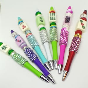 May include: A collection of six ballpoint pens featuring decorative beads and mahjong tiles at the top. Each pen has a barrel section covered in small, sparkling rhinestones in various colors including green, purple, blue, pink, and red.