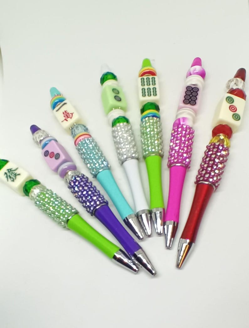 May include: A collection of six ballpoint pens featuring decorative beads and mahjong tiles at the top. Each pen has a barrel section covered in small, sparkling rhinestones in various colors including green, purple, blue, pink, and red.