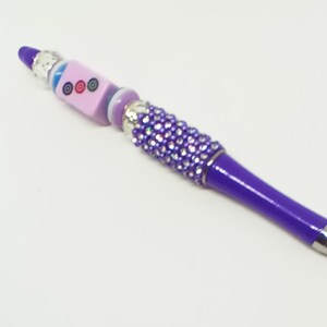 May include: A purple ballpoint pen featuring a barrel adorned with small, sparkling purple rhinestones. The top section includes a light purple cube bead with three circular designs, accented by silver-toned metal spacers and a blue bead.