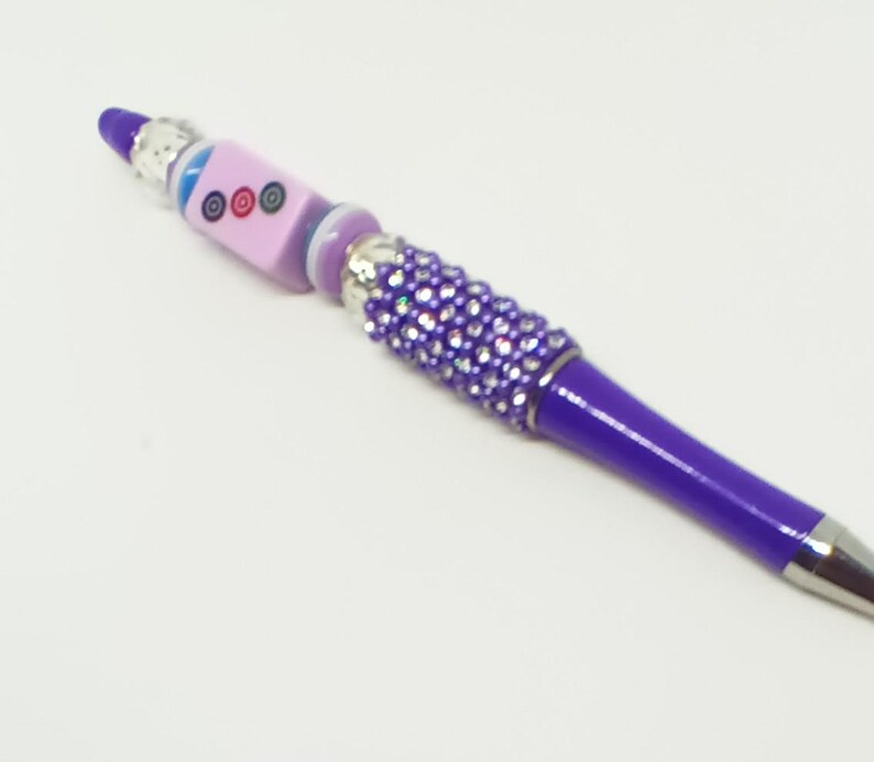 May include: A purple ballpoint pen featuring a barrel adorned with small, sparkling purple rhinestones. The top section includes a light purple cube bead with three circular designs, accented by silver-toned metal spacers and a blue bead.