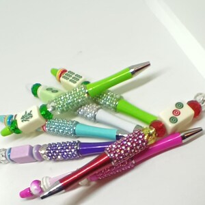 May include: A collection of ballpoint pens featuring colorful barrels, metallic tips, and decorative beads. Each pen is accented with sparkling rhinestone-covered sections and topped with various mahjong tiles, including characters and circular patterns.