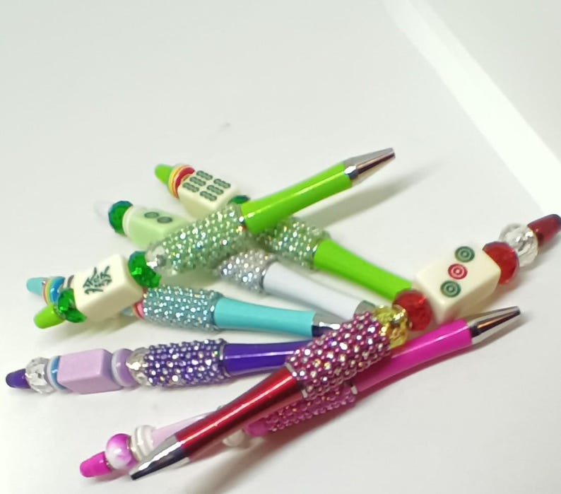 May include: A collection of ballpoint pens featuring colorful barrels, metallic tips, and decorative beads. Each pen is accented with sparkling rhinestone-covered sections and topped with various mahjong tiles, including characters and circular patterns.