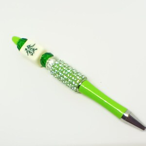 May include: A lime green ballpoint pen featuring a silver-toned metal tip and a barrel section encrusted with small, clear, sparkling rhinestones. The top of the pen is decorated with a green bead and a white mahjong tile with a black character.