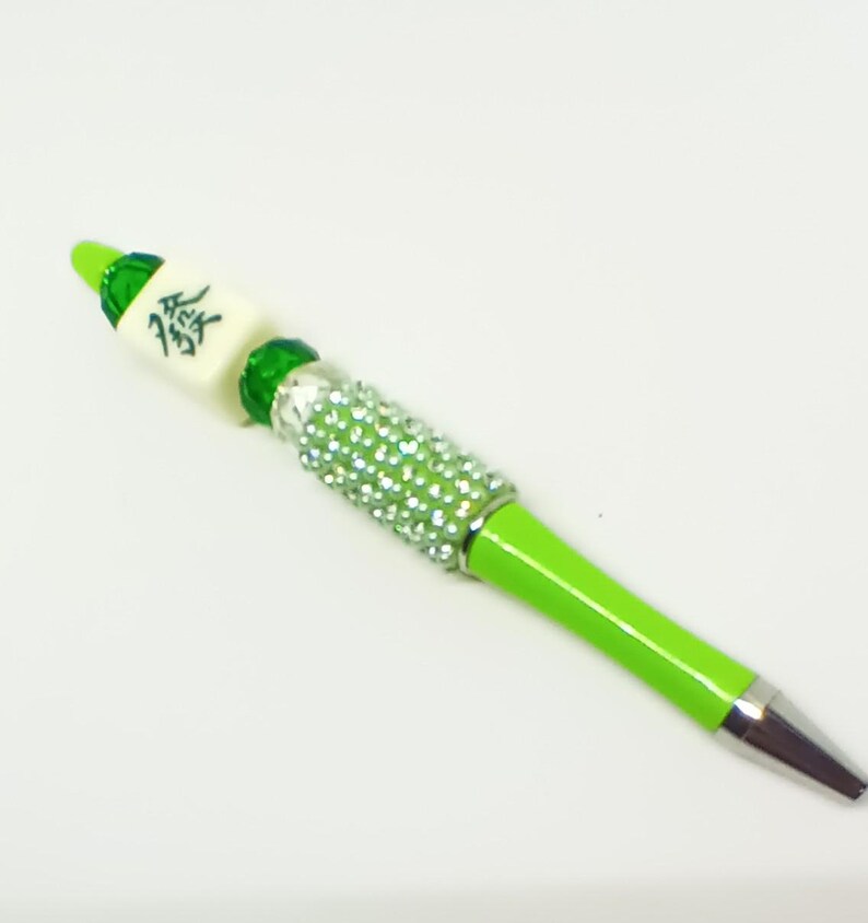 May include: A lime green ballpoint pen featuring a silver-toned metal tip and a barrel section encrusted with small, clear, sparkling rhinestones. The top of the pen is decorated with a green bead and a white mahjong tile with a black character.