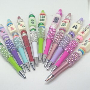 May include: A collection of ten pens featuring decorative beads and mahjong tiles. Each pen has a metallic tip and a barrel adorned with various colored rhinestones, plastic beads, and rectangular mahjong tiles with printed symbols and characters.