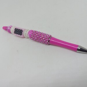 May include: A pink ballpoint pen featuring a barrel decorated with small clear rhinestones and pink beads. The upper section includes a white cylindrical bead with a black circular pattern, accented by additional pink and white decorative beads.