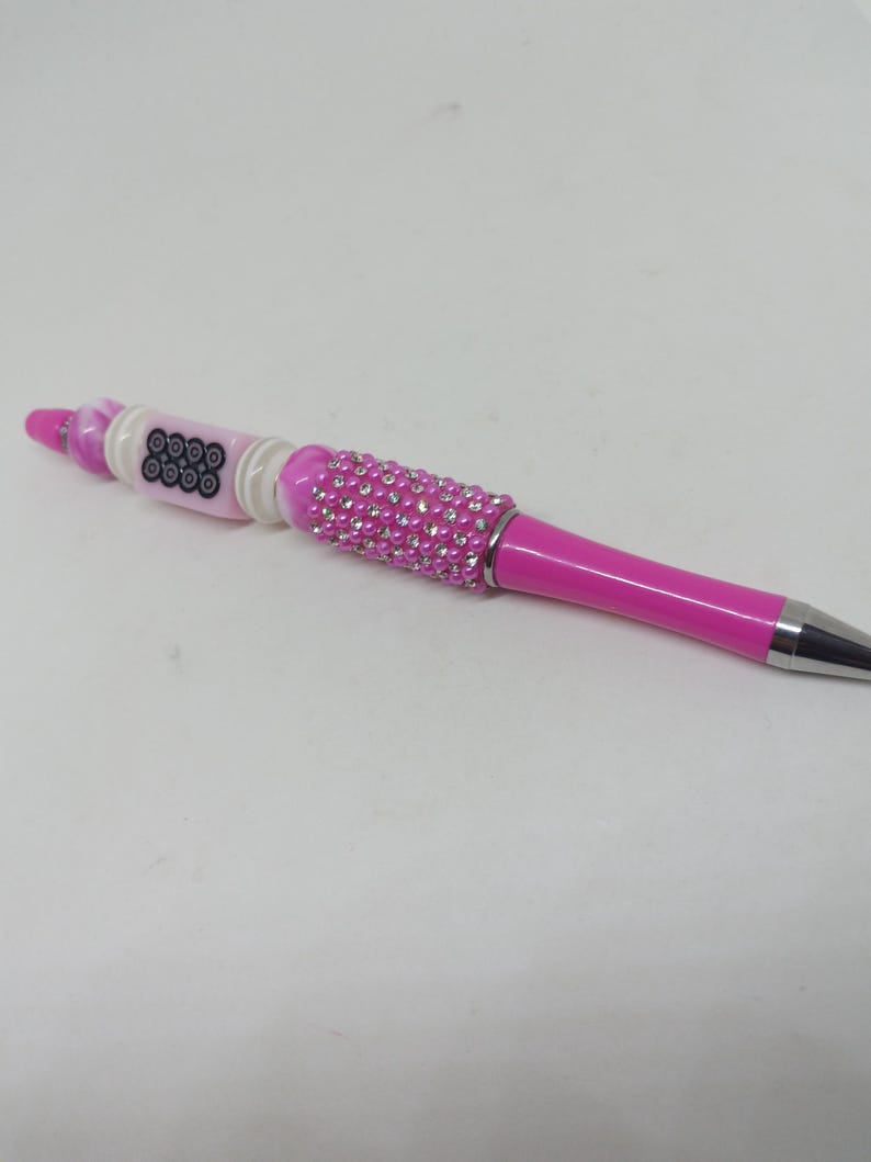 May include: A pink ballpoint pen featuring a barrel decorated with small clear rhinestones and pink beads. The upper section includes a white cylindrical bead with a black circular pattern, accented by additional pink and white decorative beads.