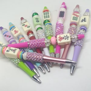 May include: A collection of pens featuring mahjong tiles as decorative toppers. Each pen barrel is adorned with colorful rhinestones and beads in various shades including pink, green, purple, and blue. The mahjong tiles display various symbols.