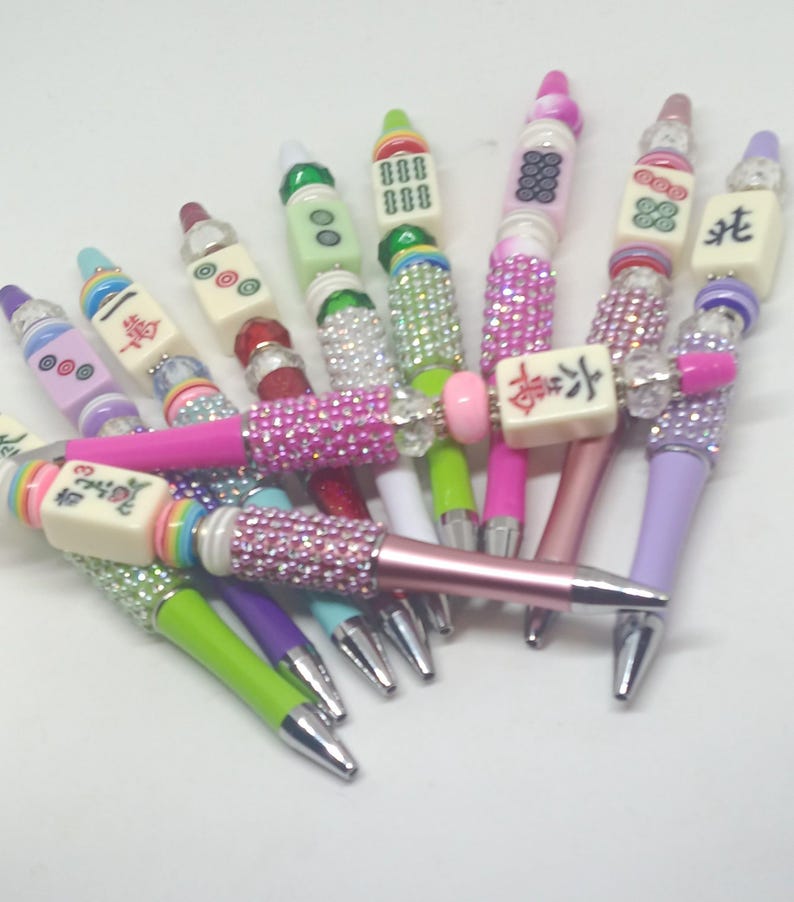 May include: A collection of pens featuring mahjong tiles as decorative toppers. Each pen barrel is adorned with colorful rhinestones and beads in various shades including pink, green, purple, and blue. The mahjong tiles display various symbols.