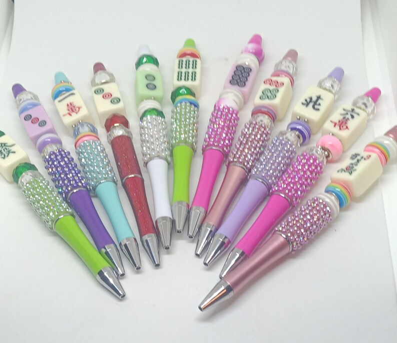 May include: A collection of ten pens featuring decorative beads and mahjong tiles. Each pen has a metallic tip and a barrel adorned with various colored rhinestones, plastic beads, and rectangular mahjong tiles with printed symbols and characters.