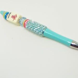 May include: A turquoise ballpoint pen featuring a silver tip and a decorative barrel adorned with a light blue crystal-encrusted section, a pink ring, a light blue bead, a cream-colored mahjong tile with red Chinese characters, and rainbow rings.