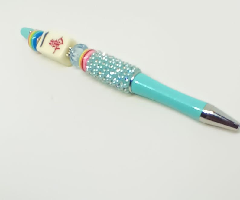May include: A turquoise ballpoint pen featuring a silver tip and a decorative barrel adorned with a light blue crystal-encrusted section, a pink ring, a light blue bead, a cream-colored mahjong tile with red Chinese characters, and rainbow rings.
