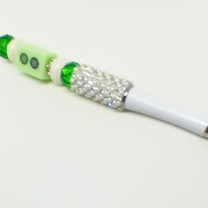 May include: A white ballpoint pen featuring a decorative barrel section covered in small silver rhinestones, accented with green faceted beads, white spacers, and a light green rectangular bead with two dark circular inlays at the top end.