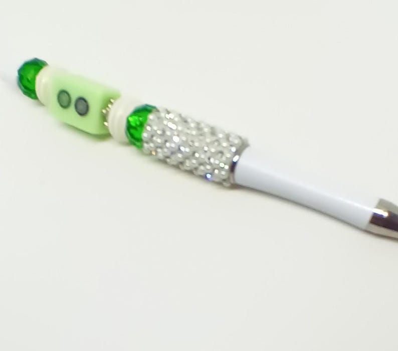 May include: A white ballpoint pen featuring a decorative barrel section covered in small silver rhinestones, accented with green faceted beads, white spacers, and a light green rectangular bead with two dark circular inlays at the top end.